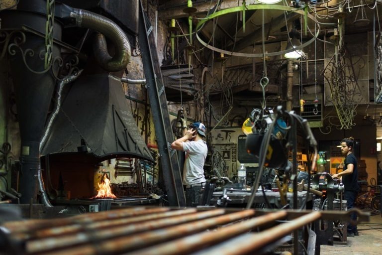 Une forge avec deux hommes au travail. L'un d'eux porte une chemise blanche et une casquette de baseball, le dos tourné, tout en parlant au téléphone. On voit la forge en activité avec des outils suspendus. Le deuxième homme, en chemise noire, est à un autre poste de travail en arrière-plan. La boutique de la Ferronnerie d'Art de Bordeaux est remplie de tiges et d'outils en métal.