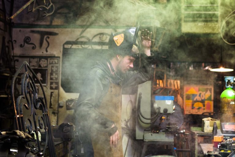 Un homme portant un casque de soudeur et un tablier de protection se tient debout dans un atelier faiblement éclairé et encombré, rempli d'outils et de machines divers. Une épaisse fumée ou de la vapeur obscurcit partiellement la scène, créant une atmosphère brumeuse. Cet artisan, absorbé par sa tâche, incarne l'essence de la Ferronnerie d'Art Bordeaux.