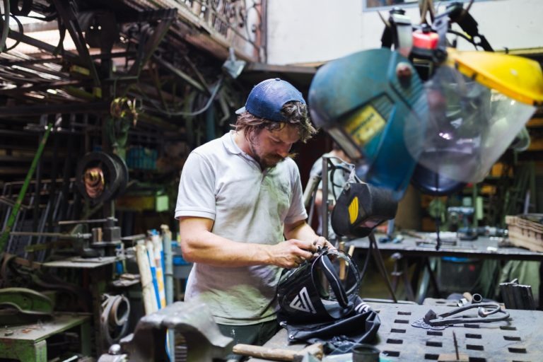 Une personne dans un atelier, vêtue d'une chemise blanche sale et d'une casquette de baseball foncée, est concentrée sur sa tâche. Autour d'elle se trouvent divers outils et machines, notamment du matériel de soudage. L'atelier encombré de la Ferronnerie d'Art Bordeaux est rempli de matériaux, d'étagères et d'un poste de travail débordant d'outils.