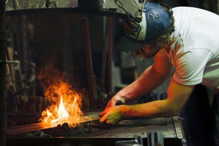 Dans l'atelier faiblement éclairé de la Ferronnerie d'Art de Bordeaux, un homme vêtu d'une chemise blanche, de gants et d'une casquette bleue travaille intensément avec une tige de métal au-dessus d'un feu vif et flamboyant. La scène capture l'effort concentré impliqué dans le travail de forge et de métal.