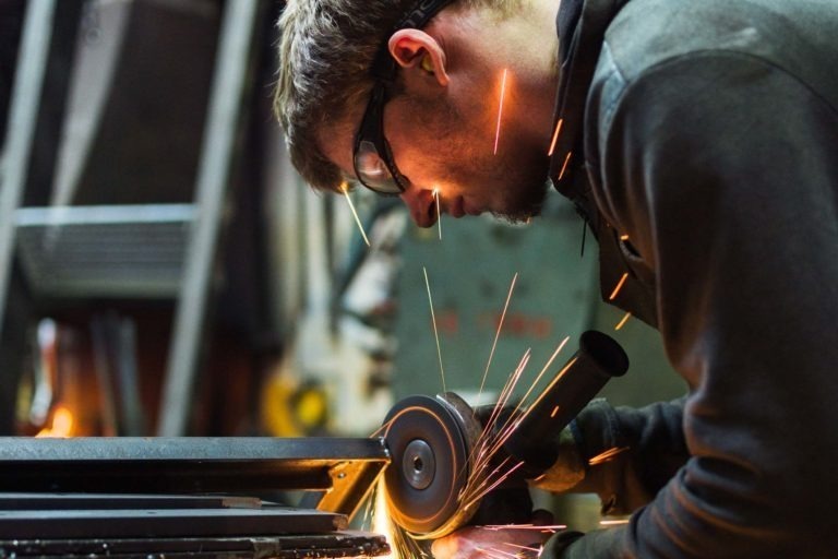 Une personne portant des lunettes de protection et des gants utilise une meuleuse d'angle, ce qui fait jaillir des étincelles brillantes. Elle est concentrée sur sa tâche, travaillant sur une pièce de métal dans un atelier de Ferronnerie d'Art de Bordeaux. Une échelle et divers équipements sont flous en arrière-plan.