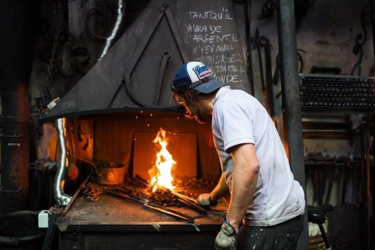 Une personne en chemise blanche et casquette travaille dans une forge, surveillant un feu ardent avec des outils de travail du métal. L'atelier de la Ferronnerie d'Art Bordeaux est rempli d'outils et d'équipements divers. Des notes manuscrites sont visibles sur une surface métallique au-dessus de la forge. Le décor est faiblement éclairé, mettant en valeur la lueur du feu.