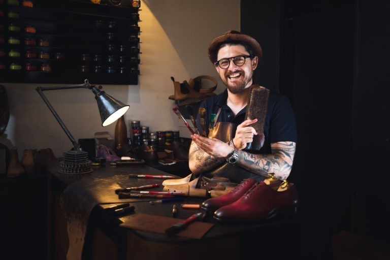 Une personne souriante, vêtue d'une chemise sombre, tatouée sur les bras, portant des lunettes et un chapeau, se tient debout dans un atelier. Elle tient des brosses et du cirage, debout derrière une table avec divers outils d'entretien des chaussures et une chaussure en cuir rouge. Des étagères avec des pots de cirage colorés sont à l'arrière-plan - le portrait créatif d'un artisan de Bordeaux et Gironde.