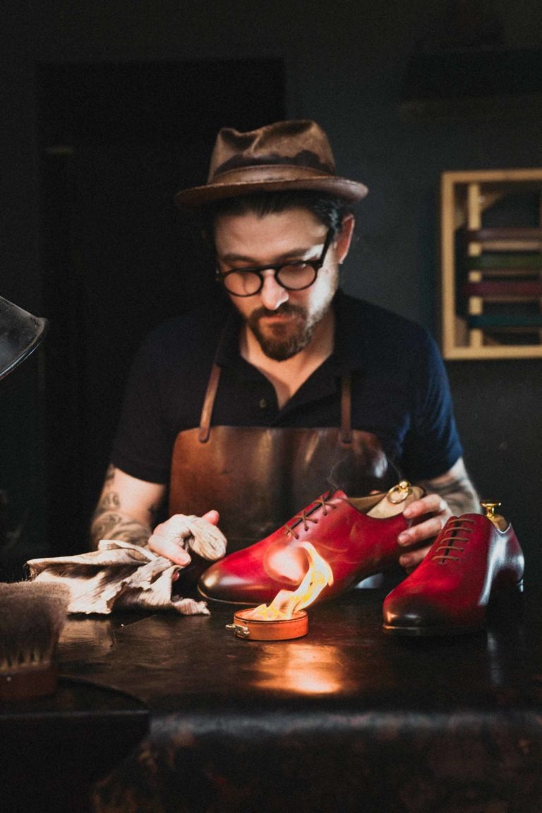 Un homme portant un chapeau marron, des lunettes et une chemise sombre se concentre sur la fabrication de chaussures en cuir rouge. Il est assis à un établi sous un éclairage tamisé, tenant une brosse à cirage pendant qu'une autre chaussure est chauffée à la flamme nue. L'espace de travail regorge d'outils et de matériaux en cuir, semblables à des portraits d'artisans de Bordeaux et de Gironde.