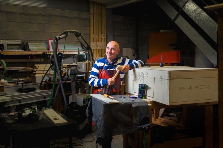 Un homme en chemise rayée travaille dans un atelier encombré d'outils et d'équipements divers. Il tient un outil en bois, travaillant peut-être sur une grande boîte en bois aux motifs dessinés. La scène ressemble à celle des portraits d'artisans de Bordeaux et de Gironde, chaleureusement éclairée pour créer une atmosphère concentrée au milieu d'équipements industriels et d'un escalier partiellement visible.