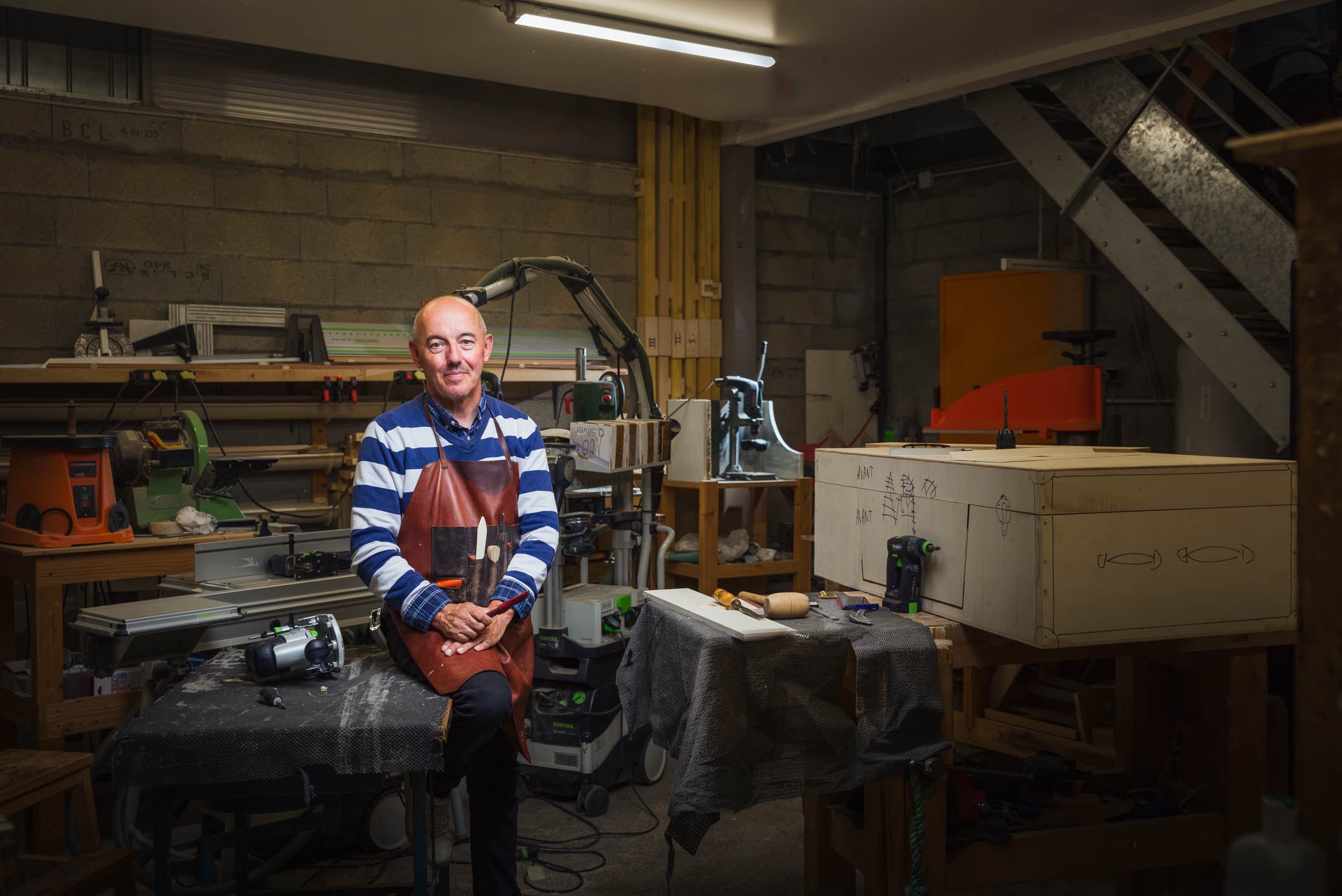 Un homme portant un tablier marron et une chemise à rayures bleues se tient debout dans un atelier de menuiserie, souriant. Il est entouré de divers outils et équipements de menuiserie, dont un grand établi avec des pièces de bois et une scie. L'atelier a un mur en béton et une atmosphère industrielle.