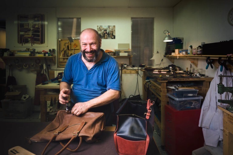 Un homme souriant en chemise bleue travaille à la fabrication d'un sac en cuir dans un atelier. L'espace de travail est encombré d'outils, de matériaux et de sacs partiellement terminés. L'arrière-plan présente des étagères et un établi remplis de divers outils de travail du cuir, avec une lampe éclairant le processus méticuleux de l'artisan de Bordeaux et de Gironde.