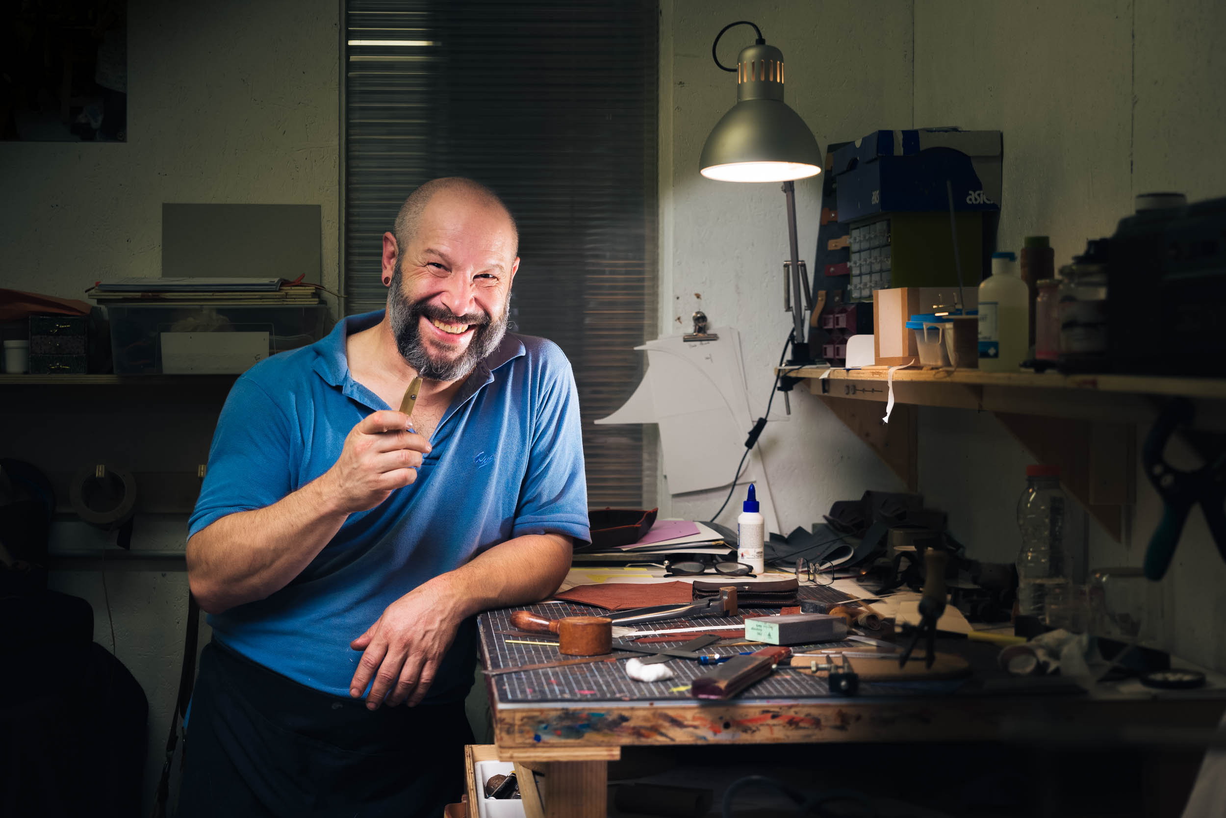 Un homme souriant, le crâne rasé et la barbe, se tient debout dans un atelier faiblement éclairé à Bordeaux. Il tient un petit outil dans sa main droite et porte un polo bleu. L'espace de travail contient divers outils, matériaux, colle et une lampe de bureau, créant un environnement encombré mais industrieux typique des artisans.