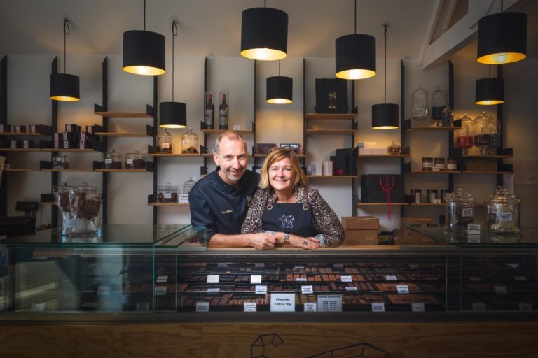 Un homme et une femme, tous deux souriants, se tiennent derrière un comptoir en verre rempli d'une gamme de chocolats. Ils se trouvent dans un magasin bien éclairé avec des lampes suspendues noires et des étagères remplies de divers produits et pots de chocolat. L'homme porte un uniforme noir et la femme porte un haut à motifs. Leurs portraits reflètent les fiers artisans qu'ils sont à Bordeaux et en Gironde.