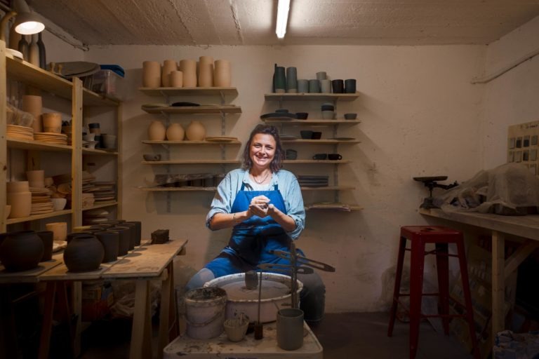 Une femme souriante, vêtue d'un tablier bleu et d'une chemise en jean, est assise à un tour de potier dans un atelier encombré. Des étagères avec divers pots en argile sont à l'arrière-plan et des outils sont éparpillés sur les tables. La pièce est bien éclairée par une seule lampe suspendue, capturant le portrait intime d'un artisan de Bordeaux et de Gironde.