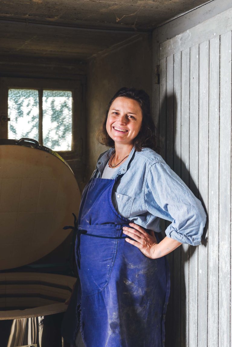 Dans une pièce rustique près de Bordeaux et de la Gironde, une femme se tient debout devant une fenêtre, vêtue d'une chemise en jean bleu et d'un tablier, souriant, la main sur la hanche. À côté d'elle, un grand objet en bois repose contre un mur en bois gris patiné. La lumière naturelle filtre à travers la fenêtre, projetant une douce lueur semblable à celle des portraits d'artisans.