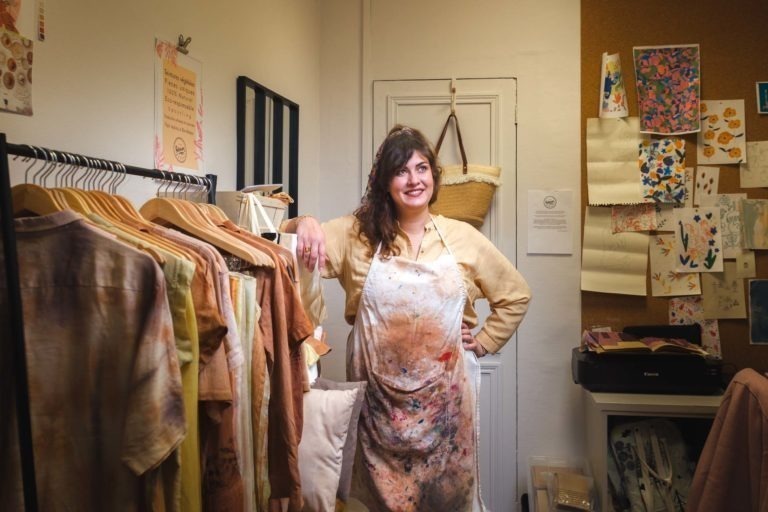 Une femme se tient debout, le sourire aux lèvres, dans une pièce lumineuse, à côté d'un portant de vêtements sur des cintres. Elle porte un tablier éclaboussé de peinture et des vêtements décontractés. Derrière elle, il y a un tableau en liège sur lequel sont épinglés divers portraits et dessins. La pièce semble être un petit atelier ou studio pour artisans de Bordeaux et Gironde.