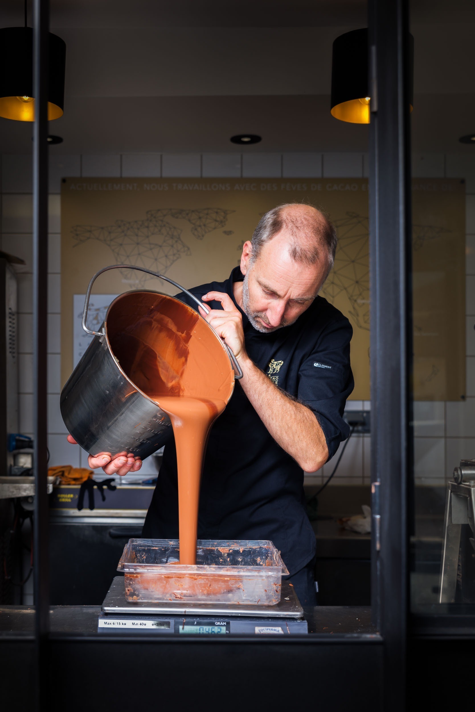 Un homme vêtu d'une veste de chef noire verse un grand récipient de chocolat fondu dans un moule rectangulaire transparent. Il se trouve dans une cuisine aux murs carrelés de blanc. Deux suspensions noires et une carte murale des origines du cacao sont visibles en arrière-plan, capturant l'essence des portraits d'artisans de Bordeaux et de Gironde. La scène est encadrée par une cloison vitrée.