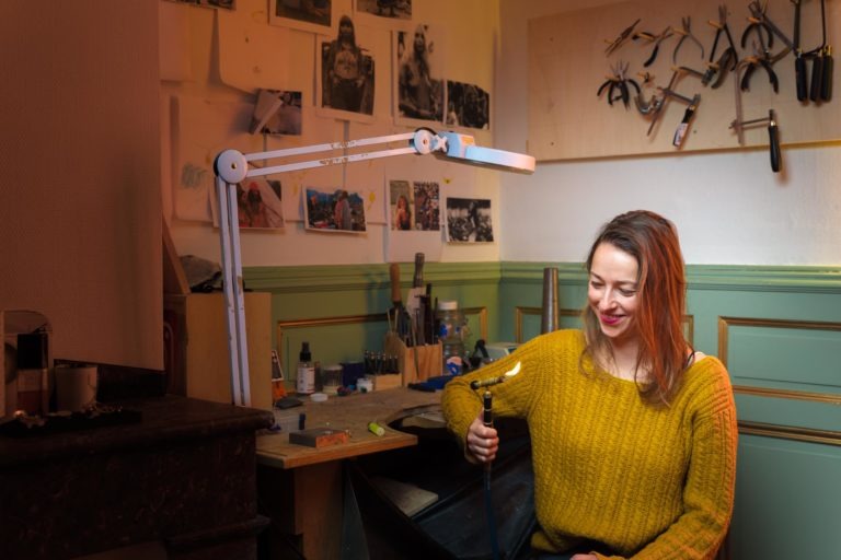 Une femme aux cheveux longs, vêtue d'un pull jaune moutarde, sourit en travaillant sur un établi d'artisan. Elle utilise un outil pour tenir un objet sous une lampe réglable et lumineuse. L'espace de travail, ressemblant à ceux des artisans de Bordeaux et de Gironde, présente des dessins, des photos et des outils sur les murs.