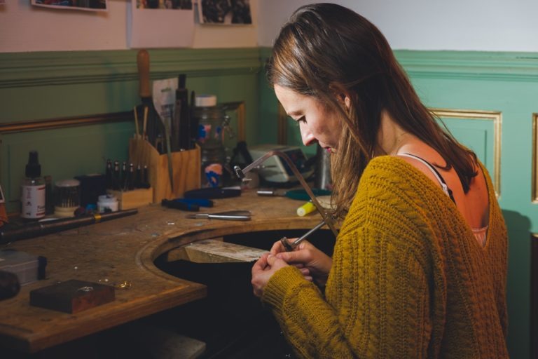 Une personne aux cheveux bruns portant un pull jaune travaille intensément sur un établi bien équipé. Des outils, des fournitures et des œuvres d'art, notamment des portraits complexes, sont visibles dans l'espace, indiquant un environnement créatif et axé sur l'artisanat. L'artisan semble utiliser une scie de bijoutier sur un morceau de tissu.