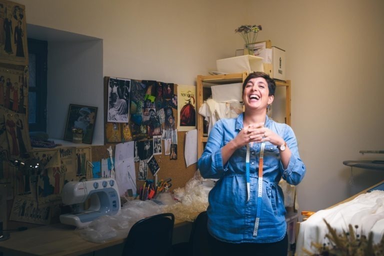 Une personne se tient dans une petite salle de couture bien éclairée à Bordeaux et en Gironde, souriant et riant. Elle porte une chemise bleue avec un mètre ruban autour du cou. La pièce contient une machine à coudre, un tableau d'affichage avec divers échantillons de tissus et des croquis de mode, ainsi que des étagères avec des matériaux et des boîtes de rangement - un véritable havre de paix pour l'artisan, parfait pour créer des portraits d'artisanat.