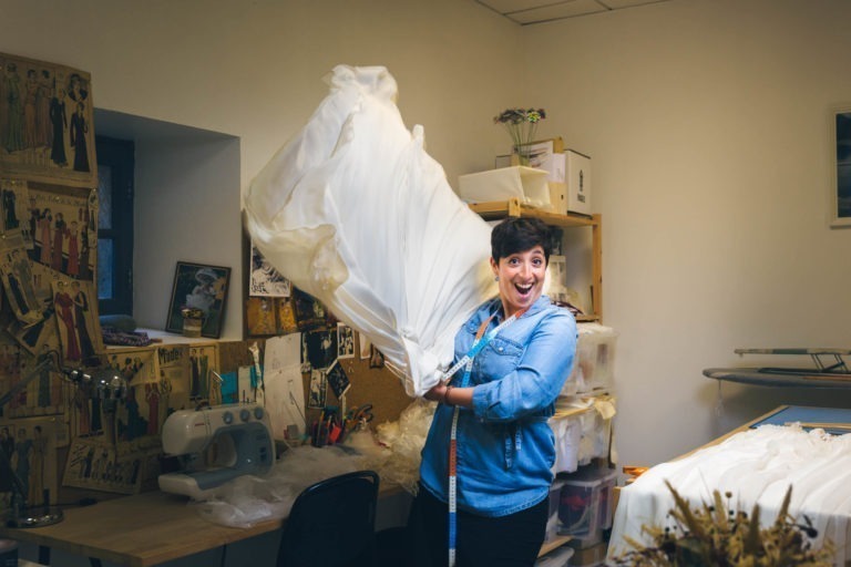 Une personne aux cheveux courts, vêtue d'une chemise en jean, se tient dans une salle de couture à Bordeaux et Gironde, tenant et agitant avec enthousiasme un grand morceau de tissu blanc. La pièce contient du matériel de couture, une machine à coudre, des illustrations colorées sur le mur et des étagères remplies de boîtes. La personne sourit.