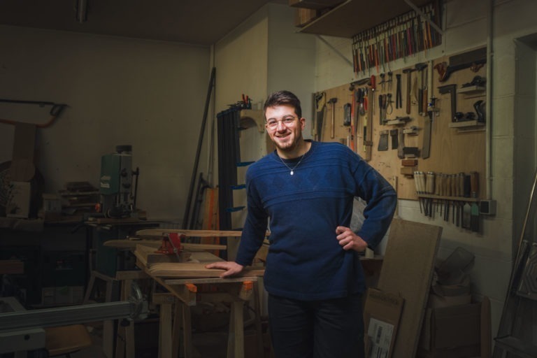 Un homme portant des lunettes et une barbe, vêtu d'un pull bleu, se tient debout, souriant, une main sur la hanche et l'autre posée sur un établi dans un atelier de menuiserie. Les portraits des artisans de Bordeaux et de Gironde ornent le mur aux côtés de divers outils, avec une machine à bois visible en arrière-plan.