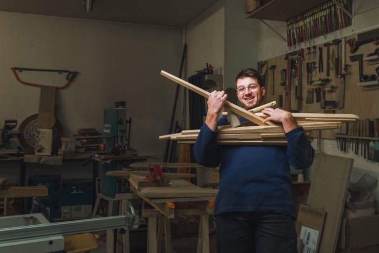 Un homme en pull bleu tient plusieurs planches de bois dans un atelier de menuiserie, semblable à celui des artisans qualifiés de Bordeaux et de la Gironde. L'atelier dispose de divers outils accrochés aux murs, notamment des scies, des marteaux et des serre-joints. Des établis et une scie à ruban sont présents à l'arrière-plan, créant un espace de travail animé et efficace.