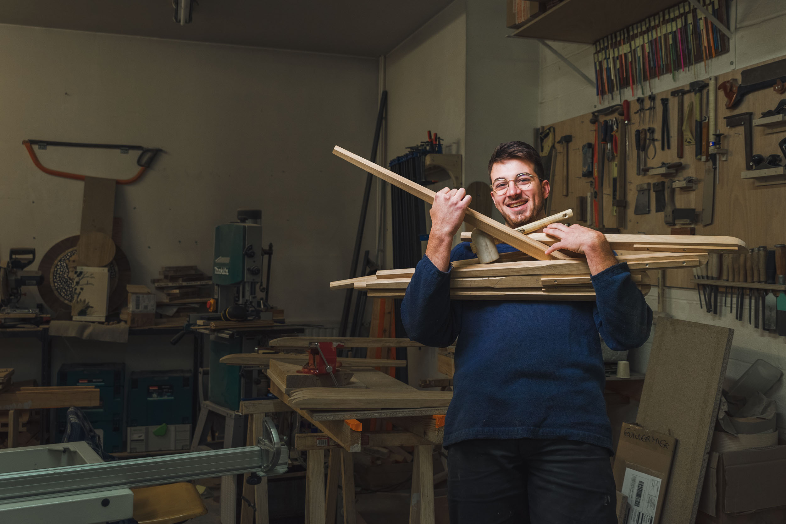 Un homme en pull bleu tient plusieurs planches de bois dans un atelier de menuiserie, semblable à celui des artisans qualifiés de Bordeaux et de la Gironde. L'atelier dispose de divers outils accrochés aux murs, notamment des scies, des marteaux et des serre-joints. Des établis et une scie à ruban sont présents à l'arrière-plan, créant un espace de travail animé et efficace.