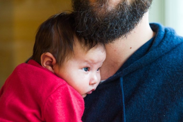 Gros plan d'un bébé appuyé sur l'épaule d'un adulte. Le bébé, vêtu d'une tenue rouge et aux cheveux noirs courts, regarde dans le vide. L'adulte, vêtu d'un pull bleu et portant une épaisse barbe noire, reste hors du cadre. La lumière d'une fenêtre les éclaire doucement, capturant des moments précieux de famille.