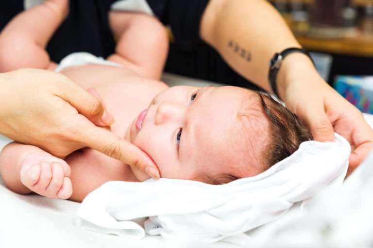 Un bébé est allongé sur une surface douce, recouvert en partie par une couverture blanche. Les mains d'un adulte touchent doucement le visage et la tête du bébé, le réconfortant. Le bébé, l'air calme, regarde vers le haut. Une partie du bras de l'adulte montre un tatouage et une montre-bracelet, capturant des moments précieux de famille.