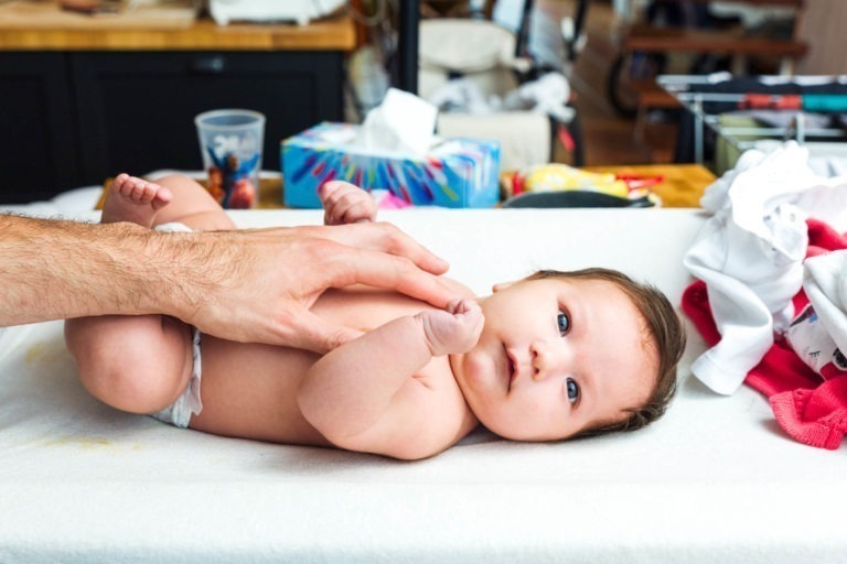 Un bébé est allongé sur un matelas à langer blanc, la main d'un homme touchant doucement son ventre, capturant des moments précieux. Le bébé, portant une couche et regardant vers le haut avec de grands yeux, se trouve dans une maison confortable et habitée. L'arrière-plan comprend une boîte de mouchoirs, une tasse et d'autres objets divers.