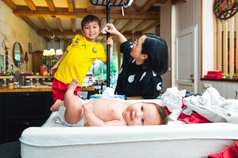 Un bébé est allongé sur une table à langer rembourrée, regardant vers la caméra. Une femme, légèrement souriante, se tient derrière elle, une main levée, semblant ajuster un mobile. Un jeune garçon en tenue jaune et rouge se tient à côté d'elle, souriant et tenant la table à langer. Capturez des moments précieux en famille dans ce cadre douillet.