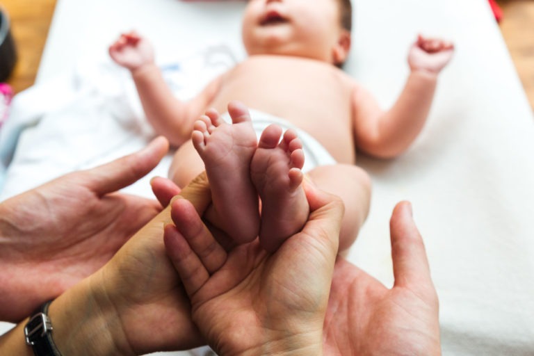 Un bébé est allongé sur le dos sur une table à langer, portant une couche. Deux paires de mains d'adultes tiennent délicatement les pieds du bébé. Les bras du bébé sont levés et son visage est flou. L'arrière-plan est légèrement flou, capturant ces moments précieux de famille avec tendresse.