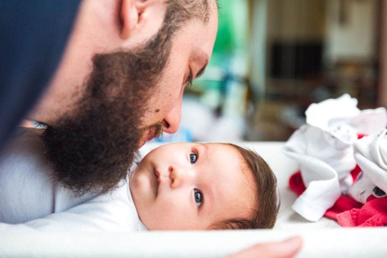 Un homme barbu se penche amoureusement vers le bébé allongé qui le regarde et l'embrasse sur le front. Le bébé, aux grands yeux curieux et vêtu d'une tenue blanche, est entouré de peluches et de vêtements, créant une atmosphère chaleureuse. L'arrière-plan est légèrement flouté pour capturer des moments précieux en famille.