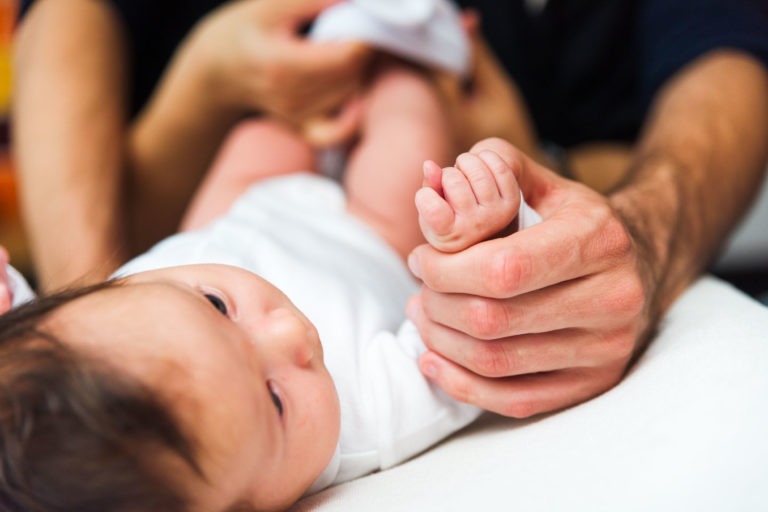 Gros plan d'un bébé allongé sur une surface blanche, vêtu d'une tenue blanche. Les mains d'un adulte tiennent délicatement le pied droit du bébé, l'autre main le soutenant. La tête du bébé est légèrement tournée sur le côté, le regard levé avec une expression calme. Capturez ces moments précieux en famille. L'arrière-plan est flou.