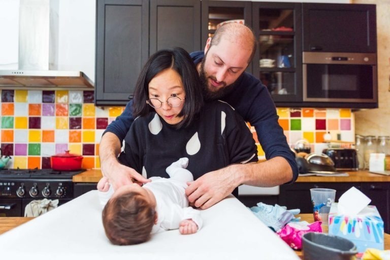 Une femme portant des lunettes et un homme s'occupent d'un bébé allongé sur une table à langer dans une cuisine aux couleurs vives, capturant de précieux moments en famille. L'arrière-plan présente un dosseret en carreaux multicolores et des armoires sombres. Divers articles pour bébé sont éparpillés sur le comptoir.