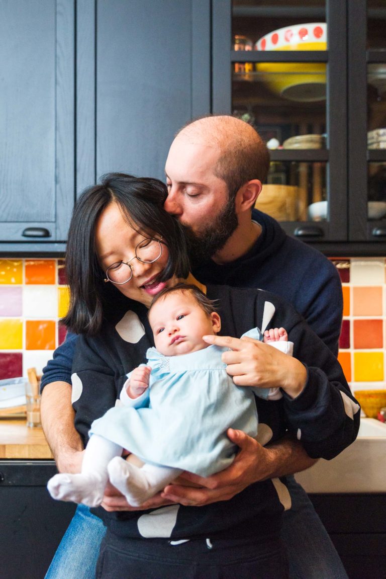 Un couple tient un bébé dans une cuisine aux placards noirs et au dosseret en carrelage coloré. La femme aux lunettes sourit tandis que l'homme barbu lui embrasse la tête. Le bébé, vêtu d'une robe bleu clair, est bercé dans leurs bras, regardant la caméra avec de grands yeux, capturant des moments précieux en famille.