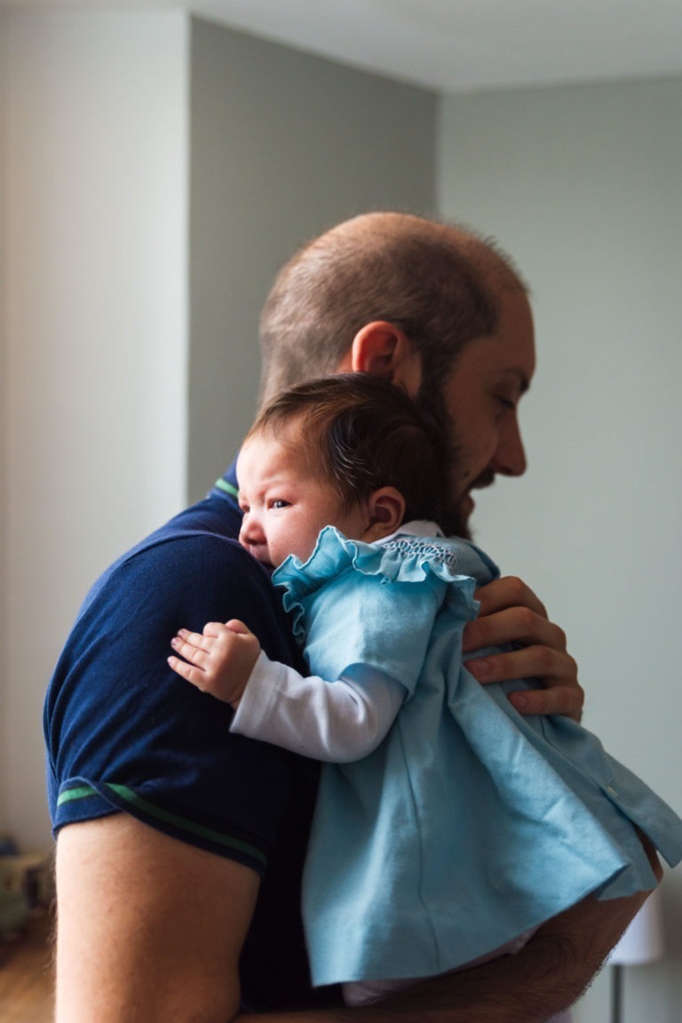 Un homme barbu tient doucement un nouveau-né contre son épaule, capturez des moments précieux. Le bébé, vêtu d'une robe bleu clair et d'une chemise blanche à manches longues, regarde sur le côté avec de grands yeux, posant sa tête près du cou de l'homme. L'arrière-plan doucement éclairé présente des murs de couleur neutre, créant une atmosphère familiale chaleureuse.