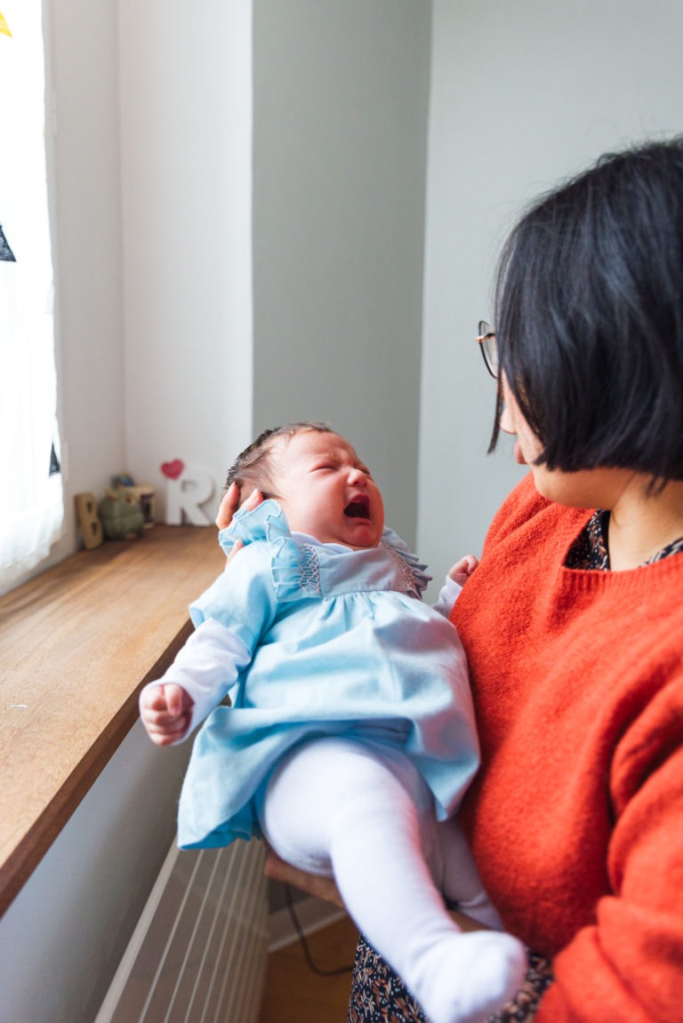 Une femme aux cheveux noirs courts et aux lunettes, vêtue d'un pull orange, tient dans ses bras un bébé en pleurs, vêtu d'une robe bleu clair et de collants blancs. Ils sont près d'une fenêtre ensoleillée avec un rebord en bois, décoré de petits ornements et d'une lettre "B" blanche, capturant de précieux moments de famille.