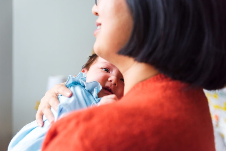 Une femme aux cheveux courts et foncés, vêtue d'un haut orange, tient un bébé dans ses bras. Le bébé, vêtu d'une tenue bleu clair, pose sa tête sur l'épaule de la femme et regarde au loin. L'arrière-plan est légèrement flou, capturant moments précieux famille mettant en valeur le moment de tendresse entre les deux.