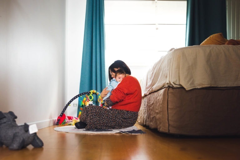 Une femme en pull rouge et jupe fleurie est assise par terre, câlinant un bébé en tenue bleue. Ils sont près d'un tapis de jeu coloré pour bébé avec des jouets, capturant des moments précieux de la famille. Derrière eux, il y a un lit avec une literie beige et de grands rideaux bleus suspendus près d'une fenêtre lumineuse. Un jouet en peluche gris est posé sur le sol à proximité.