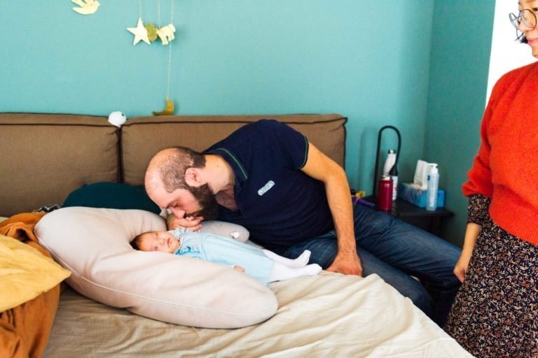 Un homme barbu est assis sur un lit, embrassant doucement un bébé allongé sur un grand oreiller, capturant de précieux moments en famille. Le bébé, habillé de bleu, repose paisiblement. À droite, une personne portant un pull rouge et une jupe à motifs se tient partiellement visible. Les murs bleu sarcelle et les décorations suspendues ajoutent du charme à la pièce.