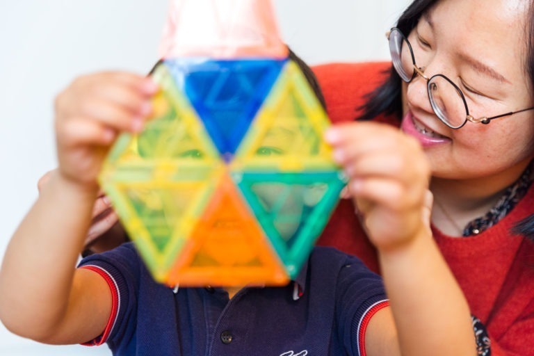 Un enfant tient un jouet géométrique coloré composé de pièces triangulaires, occultant son visage. Un adulte portant des lunettes et un pull rouge sourit et se penche près de l'enfant, le guidant doucement. Tous deux semblent engagés et heureux dans leur jeu, capturant des moments précieux en famille.