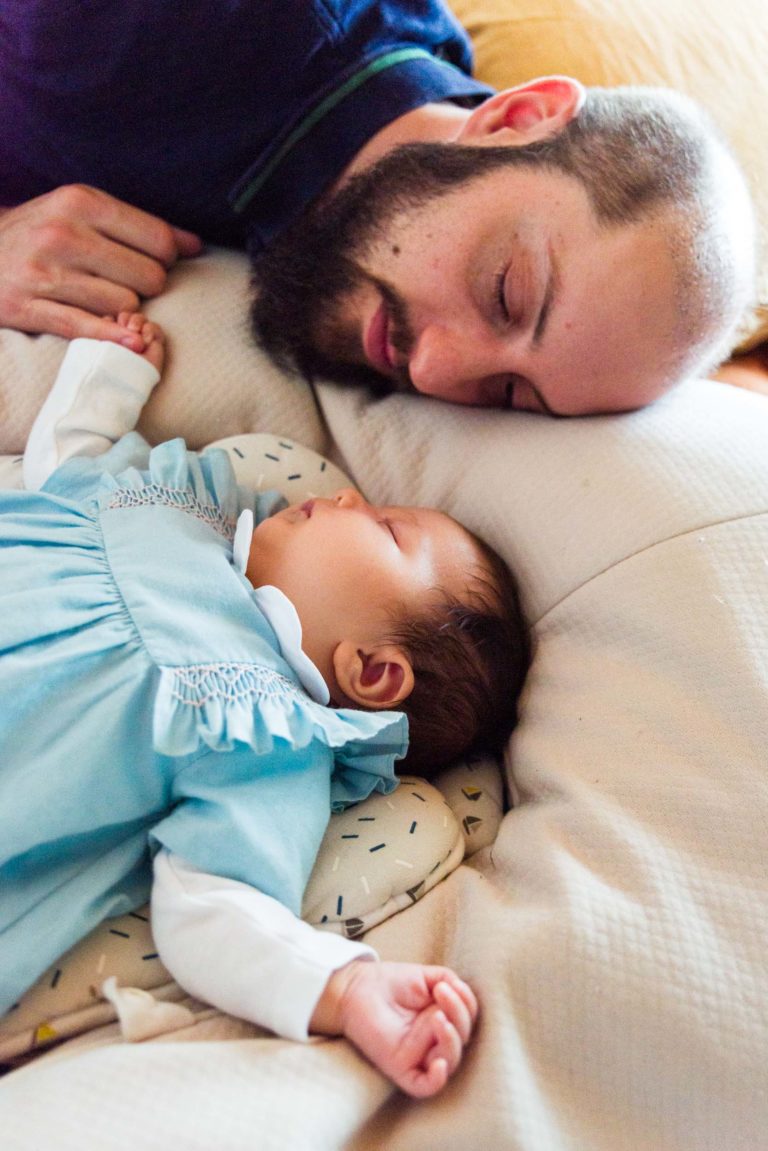 Un homme barbu est allongé sur un lit, la tête posée sur un oreiller, et regarde affectueusement un bébé endormi à côté de lui. Le bébé, vêtu d'une tenue bleu clair à volants blancs, semble paisible. Cette scène respire la chaleur et la tendresse car elle capture des moments précieux de la famille.