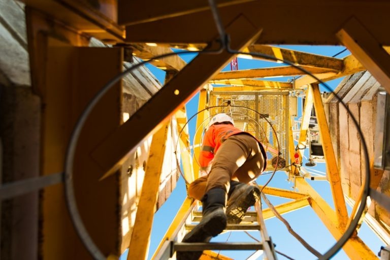 Un ouvrier de Delta Construction à Bordeaux, vêtu d'une chemise orange, d'un pantalon beige et d'un casque de sécurité blanc, grimpe sur une échelle métallique dans l'échafaudage jaune. Photographié d'en bas, le ciel bleu vif est visible à travers la structure métallique.