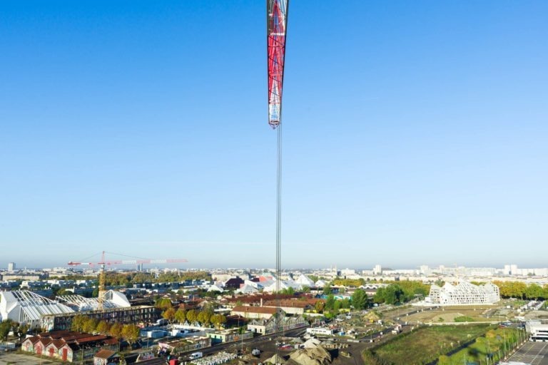 Une grande grue de chantier rouge de Delta Construction à Bordeaux domine l'horizon d'une zone urbaine composée d'un mélange de bâtiments, dont des structures blanches modernes et des bâtiments en briques plus anciens. En contrebas, un chantier de construction animé avec des équipements et des matériaux. Le ciel est clair et bleu, offrant une vue lumineuse et ensoleillée sur le paysage urbain.