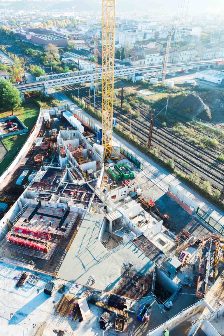 Vue aérienne d'un chantier de Delta Construction à Bordeaux avec plusieurs fondations en béton en cours de construction. Une grande grue jaune est positionnée au centre, entourée de matériaux de construction et de machines. À côté se trouvent des voies ferrées et un mélange de bâtiments, d'arbres et de routes en arrière-plan sous un ciel clair.