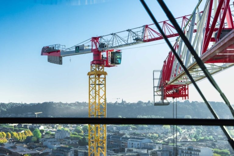 Une grande grue jaune et rouge de Delta Construction à Bordeaux est positionnée au-dessus d'un paysage urbain vu à travers une vitre. Le bras de la grue s'étend horizontalement, avec un ciel bleu clair et des collines boisées au loin en arrière-plan. Des bâtiments et des arbres sont visibles en contrebas dans le cadre urbain.