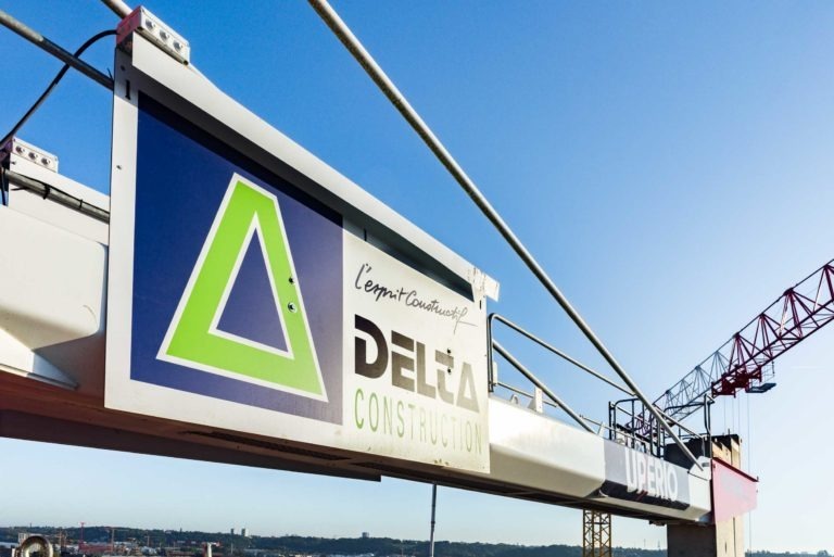 Un bras de grue de chantier sur lequel est fixé un panneau affichant le logo et le nom « DELTA CONSTRUCTION à Bordeaux » en caractères gras. Le logo présente un design triangulaire vert et bleu. En arrière-plan, un ciel bleu clair et un paysage lointain sont visibles, indiquant un chantier de construction.