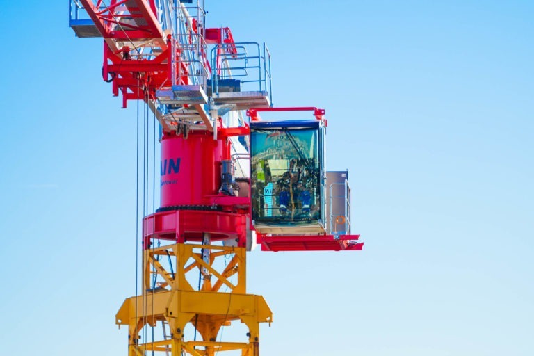 Une grue rouge et jaune de Delta Construction à Bordeaux avec un opérateur à l'intérieur d'une cabine transparente se détache sur un ciel bleu clair. La grue est équipée de divers composants de machines, de structures stabilisatrices et d'un siège conducteur avec des commandes clairement visibles. L'opérateur est vu portant un équipement de sécurité.