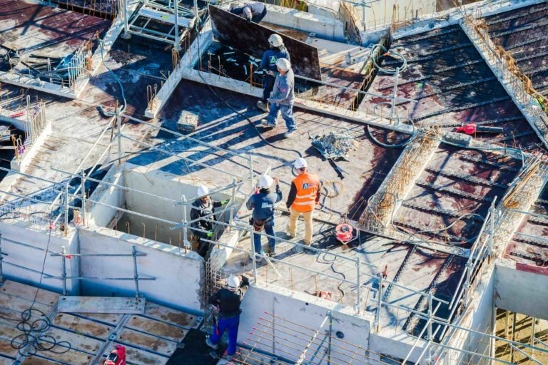 Vue aérienne d'un chantier de Delta Construction à Bordeaux sur lequel on peut voir plusieurs ouvriers en tenue de sécurité et casques. Ils sont positionnés sur une structure de bâtiment en béton, entourés de barres d'acier, de planches de bois et d'équipements de construction. Des garde-corps et des échafaudages sont visibles, et les ouvriers s'adonnent à diverses tâches.