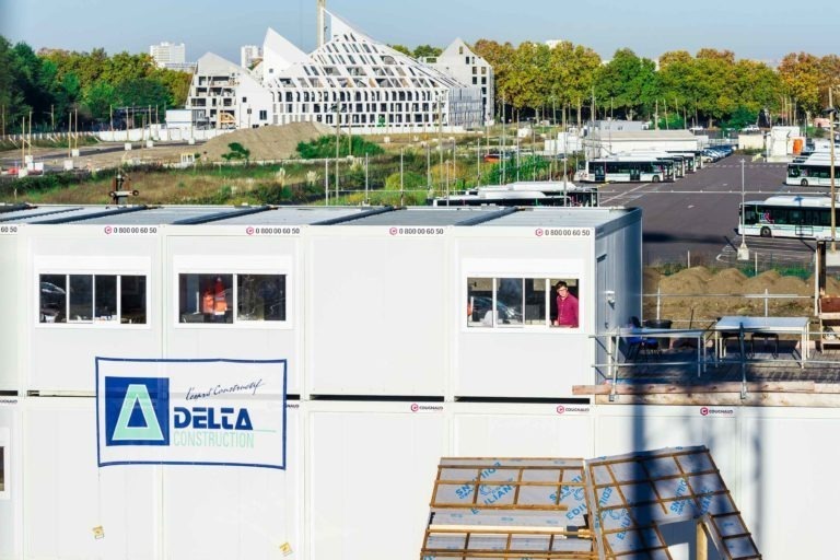 Un chantier de construction avec plusieurs bâtiments modulaires blancs de DELA Construction au premier plan, tandis que des bus stationnés et un bâtiment moderne au design triangulaire s'élèvent à l'arrière-plan. Des arbres au feuillage vert et jaune sont visibles au-delà du chantier sous un ciel dégagé, conjuguant le charme de Delta Construction à Bordeaux.