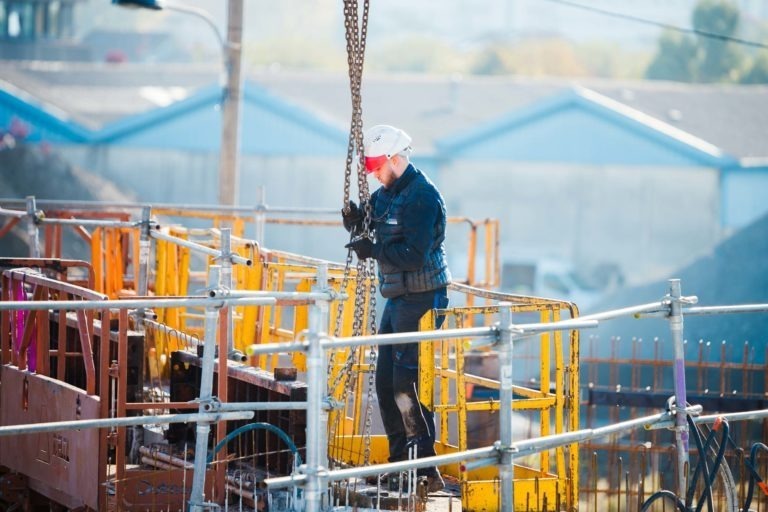 Un ouvrier du bâtiment portant un casque de chantier blanc, une veste sombre et un jean manœuvre une lourde chaîne sur le chantier de Delta Construction à Bordeaux. La zone est entourée d'échafaudages jaunes et rouges et de barrières de sécurité. Des bâtiments industriels sont visibles en arrière-plan sous un ciel dégagé.