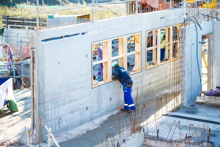 Un ouvrier de Delta Construction en tenue bleue et noire construit une structure en béton percée de fenêtres à Bordeaux. Le chantier, rempli de matériaux de construction, d'échafaudages et de barres d'armature, est éclairé par la lumière du soleil. Concentré sur sa tâche, l'ouvrier utilise des outils pour ajuster la structure en béton.